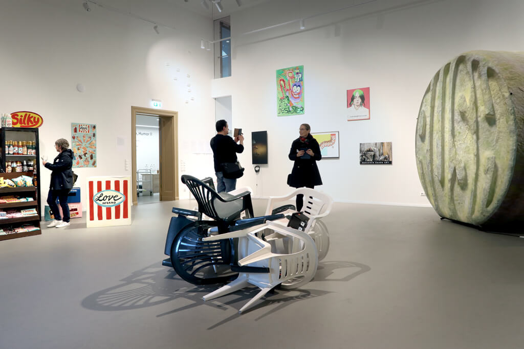 Mehrere weiße und schwarze Kunststoffstühle liegen übereinander auf dem Boden eines Museumsraums mit Kunstwerken an den Wänden und einer großen ovalen Skulptur rechts.