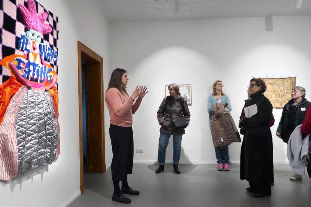 Fünf Personen stehen in einem weißen Ausstellungsraum, eine Frau in rosa Pullover spricht, an der Wand links ein farbiges, textiles Kunstwerk