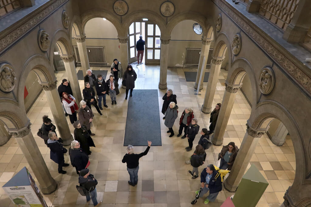 Gruppe von Menschen steht in einem großen, historischen Foyer mit Bögen und Säulen, eine Person gestikuliert in der Mitte.