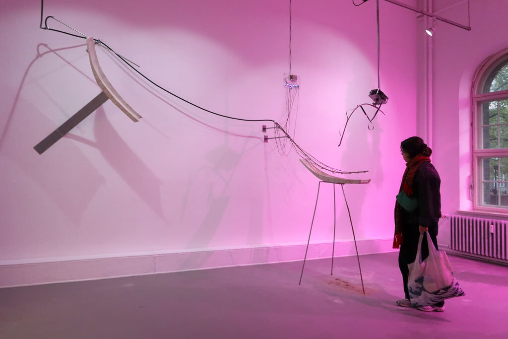 Eine Person mit Tasche betrachtet eine abstrakte, gebogene Skulptur aus Holz und Metall an einer rosa beleuchteten Wand in einem Raum mit großem Fenster.
