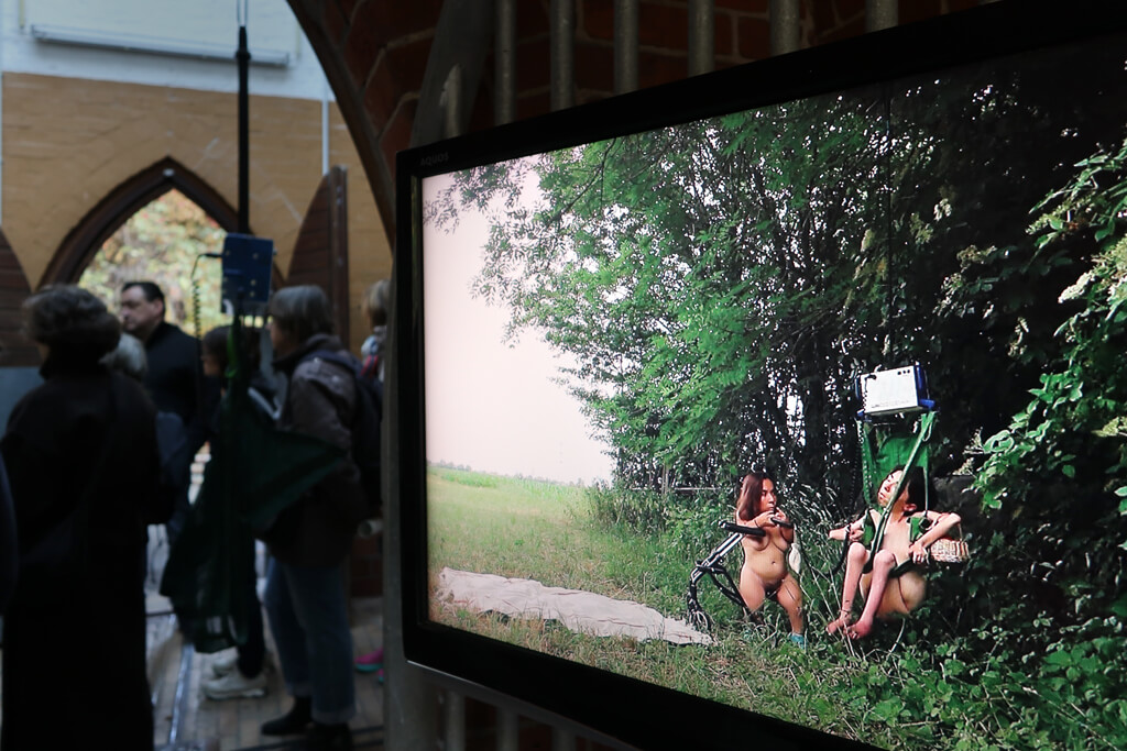 Ein Bildschirm zeigt zwei nackte Personen, die am Feldrand sitzen und von dichtem Grün und Büschen umgeben sind.