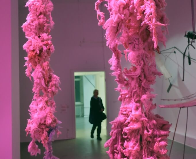 Zwei große, unregelmäßig geformte, rosa hängende Skulpturen in einem Ausstellungsraum, im Hintergrund eine Person im Türrahmen.