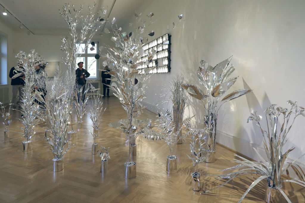 Mehrere skulpturale Pflanzen aus silberfarbenem Material stehen in einem Ausstellungsraum mit Parkettboden, im Hintergrund sind vier Personen zu sehen.