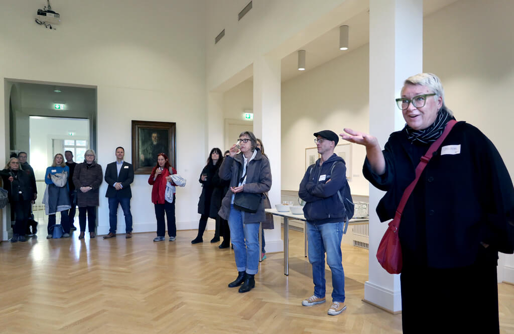 Gruppe von stehenden Menschen in einem hellen Raum mit Parkettboden, eine Person mit roter Tasche gestikuliert mit der Hand.