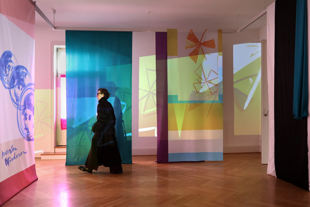 Person in schwarzem Mantel und Sonnenbrille geht durch Raum mit hängenden künstlerischen Arbeiten auf Stoffbahnen und Holzboden.