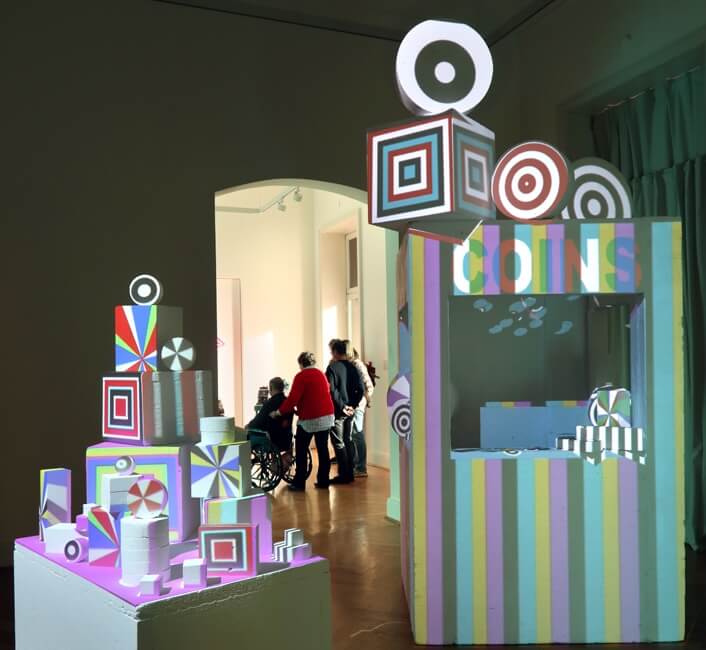 Bunte, geometrische Skulpturen mit projizierten Kreismustern und Streifen in einem Ausstellungsraum, im Hintergrund Menschen, eine Person im Rollstuhl.