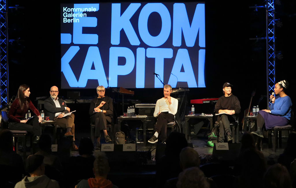 Sechs Personen sitzen auf einer Bühne vor einer großen Leinwand mit dem Text 'Kommunale Galerien Berlin' und einem teilweise sichtbarem Schriftzug