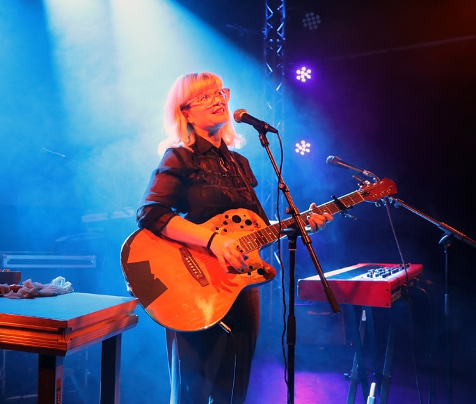 Person mit hellen Haaren spielt akustische Gitarre auf einer Bühne mit Mikrofon und Keyboard im Hintergrund.