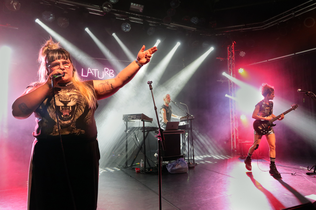 Drei Musiker auf Bühne mit Bühnenbeleuchtung, Sängerin mit Mikrofon und T-Shirt mit Löwenmotiv, Keyboarder und Gitarristin in Bewegung.
