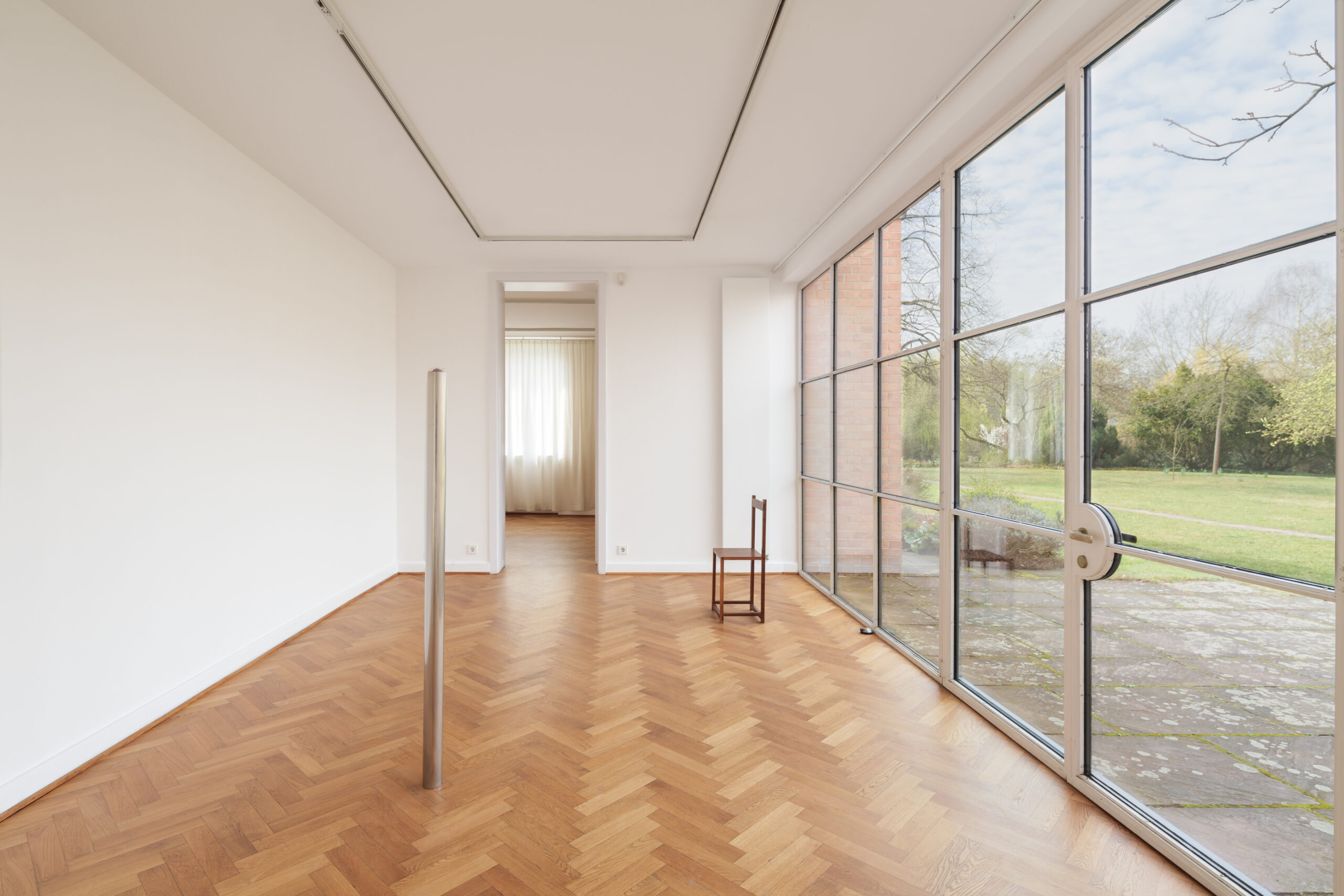 Ausstellungsraum mit einem stehenden runden Metallstab und einem großen Fenster zur Wiese.